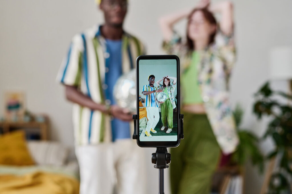 The picture shows two young vloggers. They can be seen out of focus in the background. In the foreground is a smartphone camera on a tripod. We see on the display how the two are recording a video of themselves.