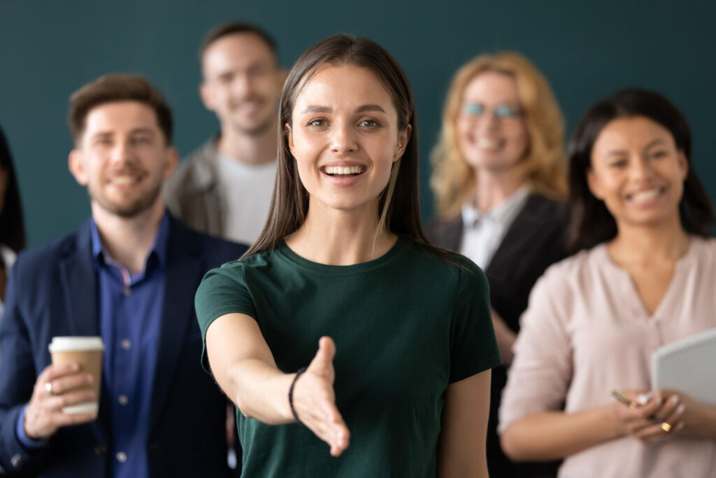 The photo shows a group of 5 young adults in the business environment. The four people in the background are out of focus, while the person in front is in focus. This young woman extends a hand invitingly to greet the photographer.