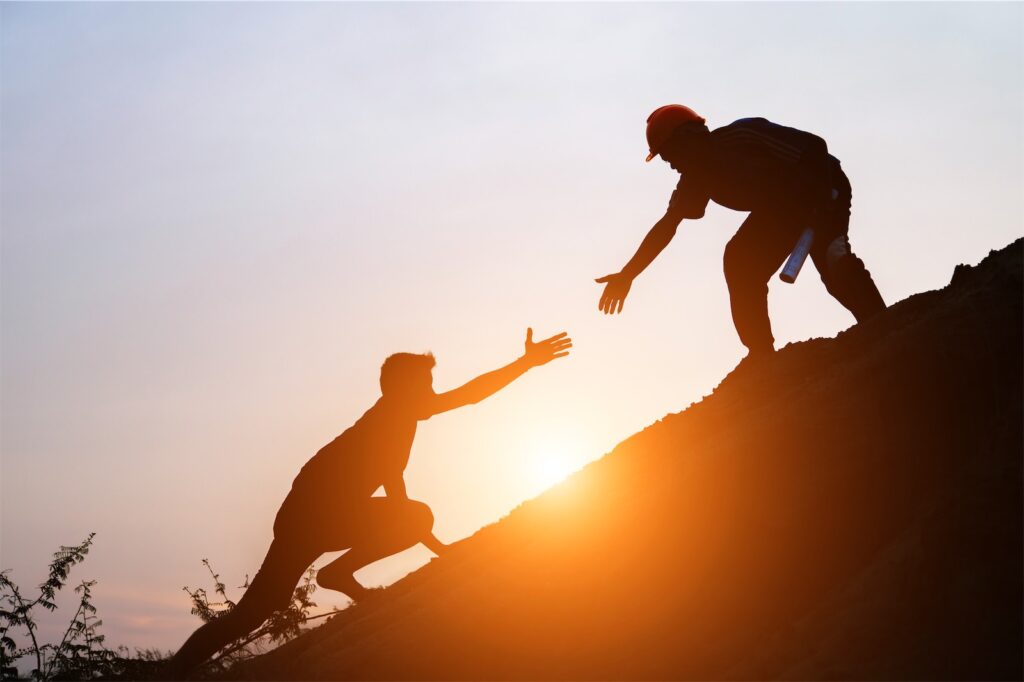 The picture shows two people on a steep hill in the backlight of a sunset. One helps the other by giving him his hand. This is a symbolic image for Individual coaching.